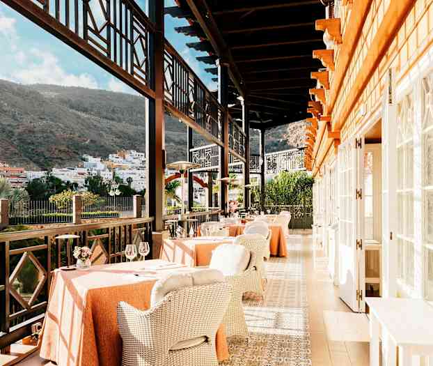 En restaurantterrasse med bord og stoler, omgitt av fjell og en by i bakgrunnen. (AI-generert tekst)