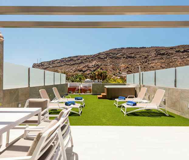 En utendørsterrasse med solsenger på kunstgress og en jacuzzi, omgitt av glassbarrierer og fjell i bakgrunnen. (AI-generert tekst)