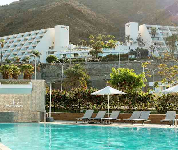 Et svømmebasseng med solsenger og parasoller, med et hvitt feriested og fjell i bakgrunnen. (AI-generert tekst)