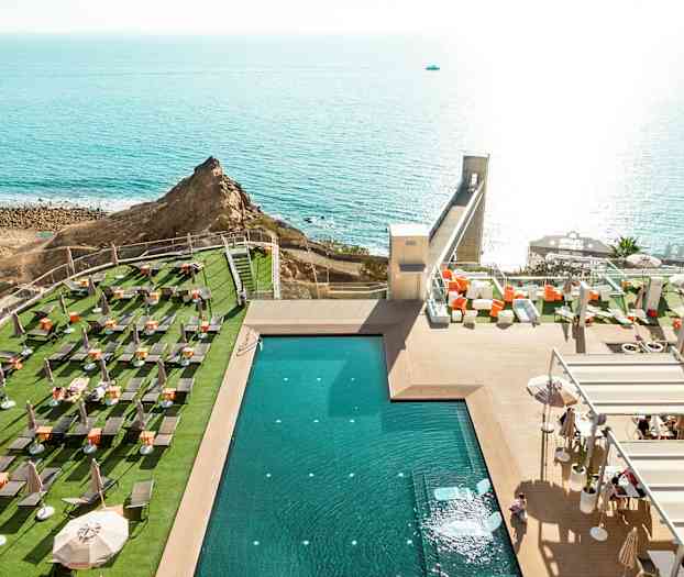 Utsikt over et svømmebasseng ved en utendørsterrasse med solsenger og havet i bakgrunnen. (AI-generert tekst)