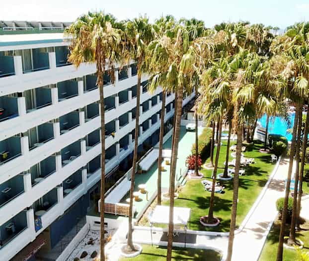 Hotell med hvite fasader omgitt av plen med palmer og et basseng. (AI-generert tekst)