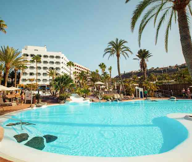 Et svømmebasseng omgitt av palmetrær og et hotell i bakgrunnen. (AI-generert tekst)