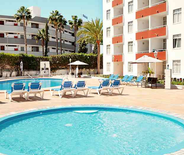 Et utendørsbasseng omgitt av solsenger, med en bygning i bakgrunnen. (AI-generert tekst)