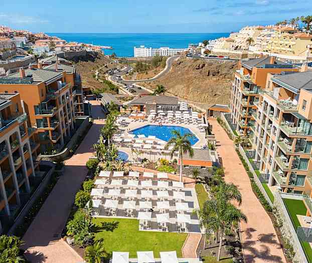 Hotellområde med basseng, solsenger og omkringliggende bygninger, med havet i bakgrunnen. (AI-generert tekst)