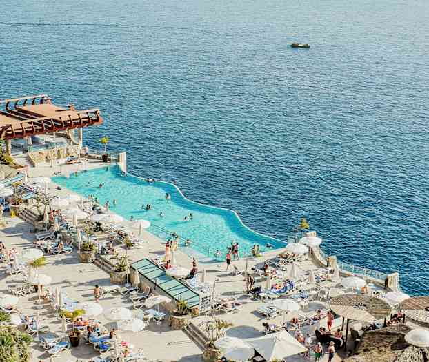 Bilde av et basseng nær stranden med solsenger og parasoller rundt, med havet i bakgrunnen. (AI-generert tekst)