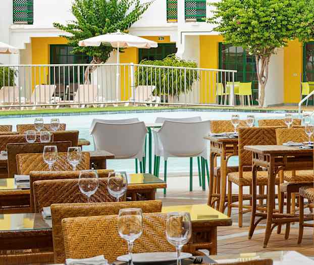 Uteservering med flettede stoler og glassbord ved et basseng. (AI-generert tekst)