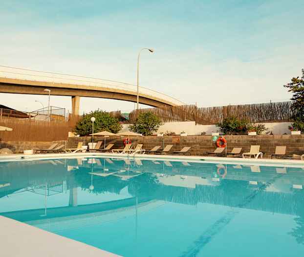 Utendørs basseng med solsenger og parasoller, omgitt av planter og mur. (AI-generert tekst)