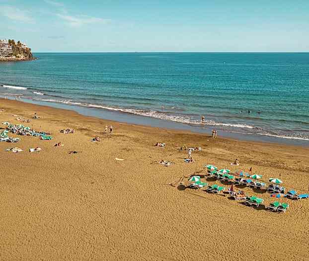 Bildet viser en sandstrand ved havet med solsenger og parasoller samt mennesker som nyter stranden. (AI-generert tekst)