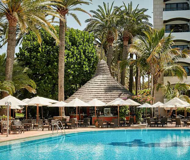 Uteområde med parasoller foran et basseng og en bygning med palmer i bakgrunnen. (AI-generert tekst)