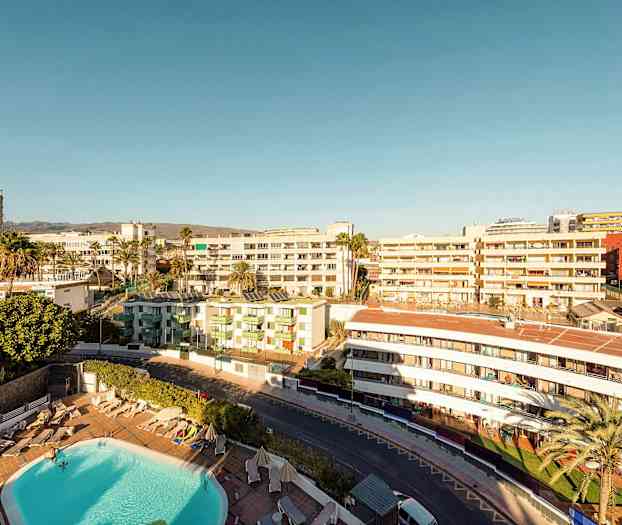 Utsikt over et hotellområde med basseng, omgitt av bygninger under en klar himmel. (AI-generert tekst)