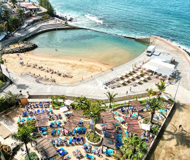 Flyfoto av en strand med solsenger og parasoller, omgitt av palmer og en gangvei. (AI-generert tekst)