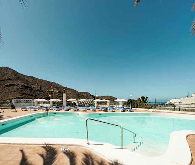 Utendørsbasseng med solsenger og parasoller, fjell i bakgrunnen og klar blå himmel. (AI-generert tekst)