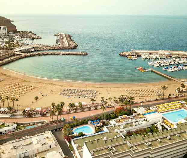 Luftfoto av en strand med parasoller og solsenger, en omkringliggende marina med båter, og nærliggende bygninger. (AI-generert tekst)