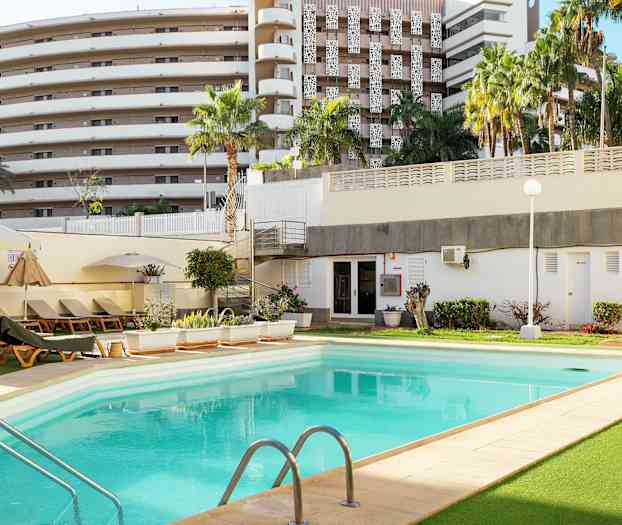 Et utendørs basseng omgitt av solsenger, grønne områder og en bygning i bakgrunnen. (AI-generert tekst)