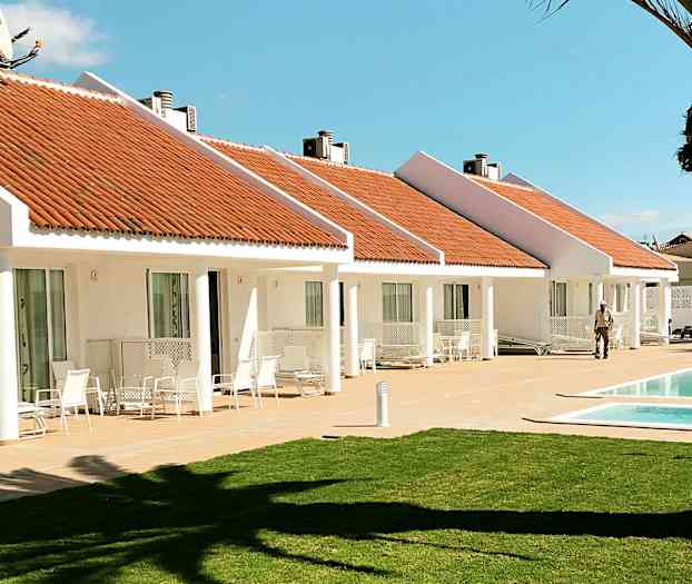 Hvite bungalower med oransje tak og et basseng foran. En plen i forgrunnen. (AI-generert tekst)