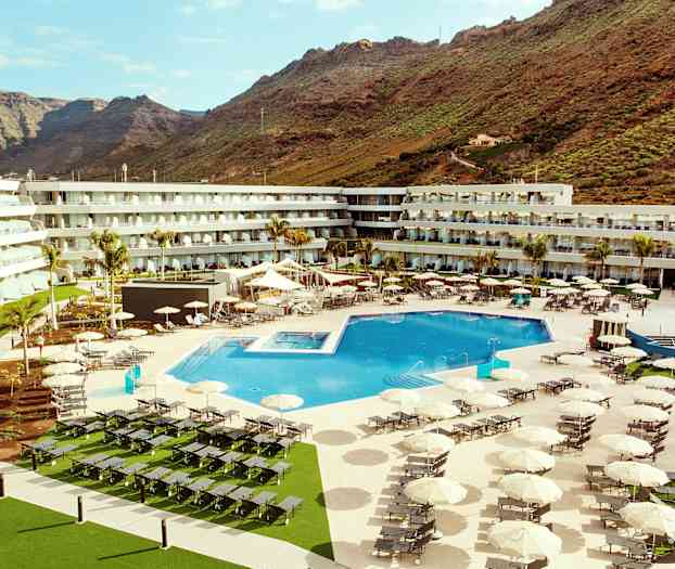 Et hotellområde med et stort utendørsbasseng, solsenger og parasoller, omgitt av bygninger og fjell i bakgrunnen. (AI-generert tekst)