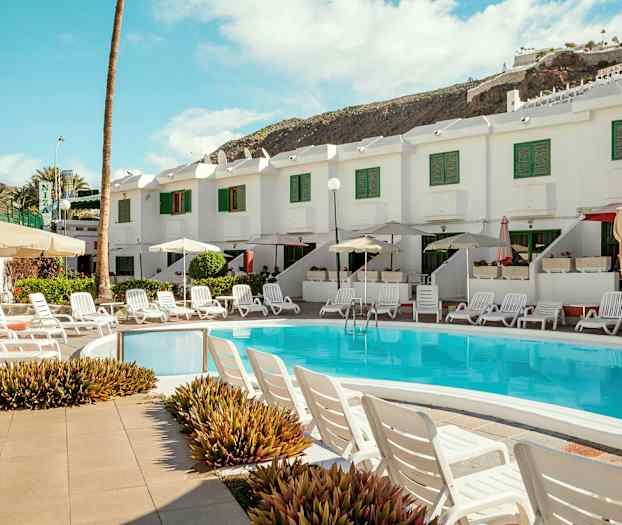 Bassengområde med solsenger og parasoller foran et hvitt hus med grønne skodder. (AI-generert tekst)