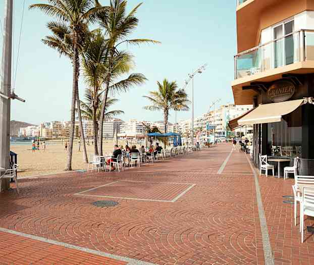 En strandpromenade med palmer, kaféstoler og bygninger i bakgrunnen. (AI-generert tekst)