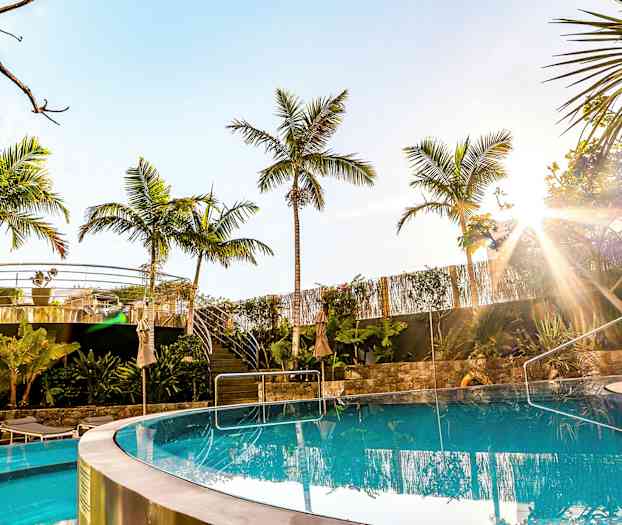 Et klart basseng med palmer og uteservering i bakgrunnen under en solrik himmel. (AI-generert tekst)