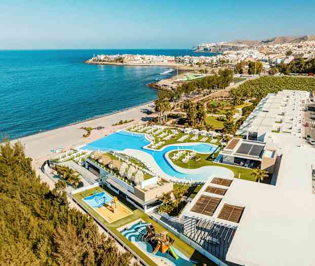 Luftfoto av et kystanlegg med bassengområde og bygninger, ved siden av en sandstrand og havet. (AI-generert tekst)