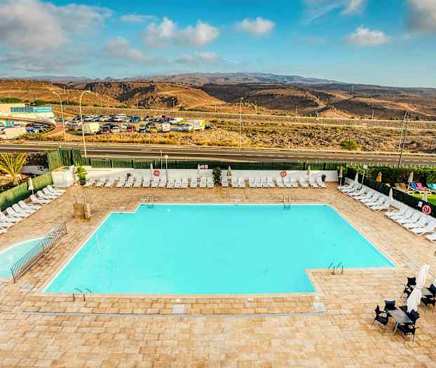 Et stort utendørs svømmebasseng omgitt av solsenger og parasoller. I bakgrunnen er en vei og et landskap med åser. (AI-generert tekst)