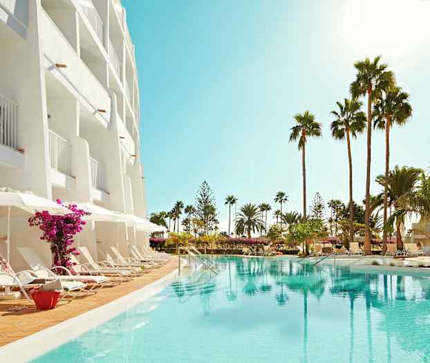 Hotellfasade med balkonger, solsenger ved bassenget og palmer i omgivelsene. (AI-generert tekst)