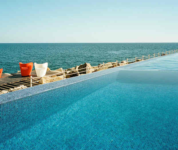 Et evighetsbasseng med utsikt over et rolig hav og et tregulv med fargerike stoler. (AI-generert tekst)