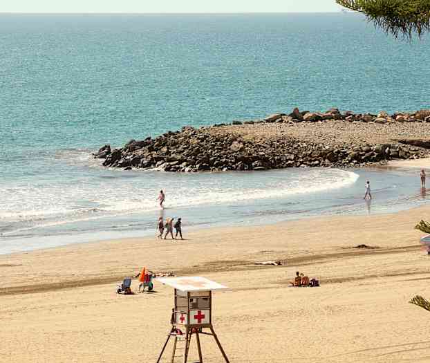 Strand med badegjester, livredningstårn og klipper ved sjøen. (AI-generert tekst)