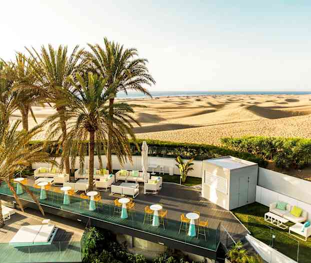En terrasse med sitteplasser og parasoller foran en ørken med sanddyner og palmer. (AI-generert tekst)