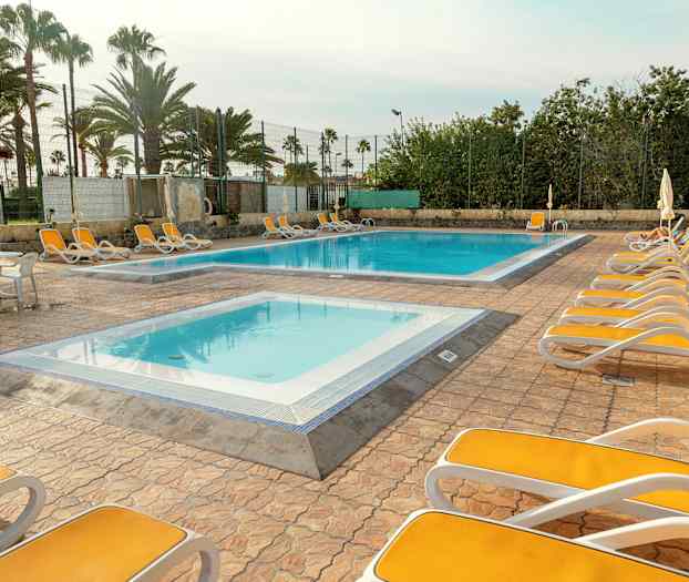 Utendørsområde med solsenger rundt to bassenger, omgitt av palmer og gjerder. (AI-generert tekst)