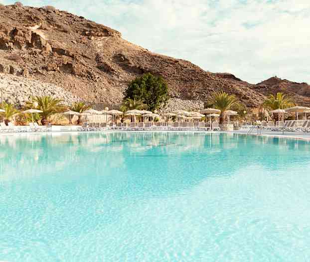 Stor basseng med solsenger og parasoller ved foten av fjell. (AI-generert tekst)
