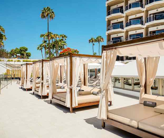 Bilde av solsenger med gardiner ved et hotellbasseng, med palmer og en bygning i bakgrunnen. (AI-generert tekst)