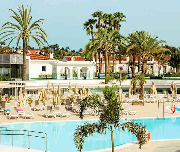 Bassengområde ved et hotell med solsenger og parasoller, omgitt av palmer og bygninger. (AI-generert tekst)