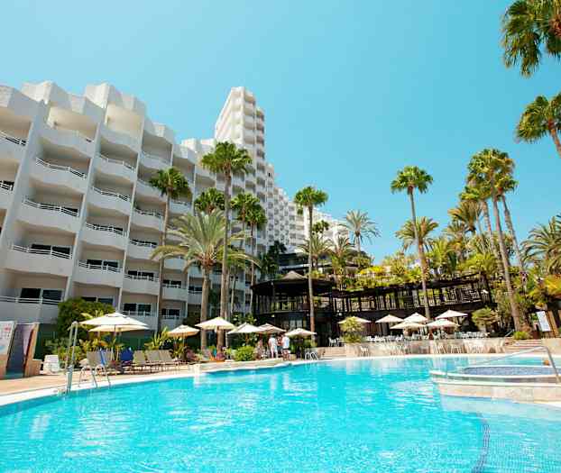 Hotellbygning med hvit fasade og et stort utendørs svømmebasseng omgitt av solsenger og parasoller. (AI-generert tekst)