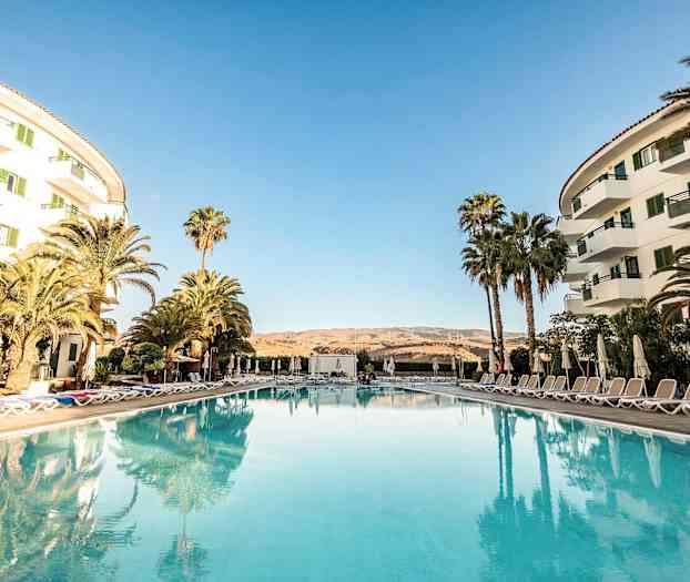 Et hotellområde med basseng i forgrunnen, omgitt av solsenger og parasoller samt omgivende bygninger på sidene. (AI-generert tekst)