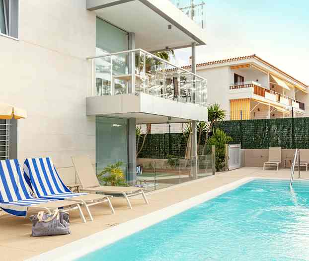 Et utendørs svømmebasseng ved et moderne leilighetskompleks med hvit fasade og balkonger. Stoler og solsenger er plassert rundt bassengområdet, og en veske ligger på en av solsengene. (AI-generert tekst)