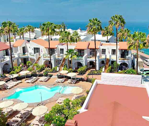 Utsikt over et hotellområde med hvitkalkede bygninger, et basseng og frodige hager, havet er synlig i bakgrunnen. (AI-generert tekst)