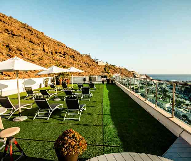 Bilde av en terrasse med solsenger, parasoller og utsikt over havet og omkringliggende klipper. (AI-generert tekst)