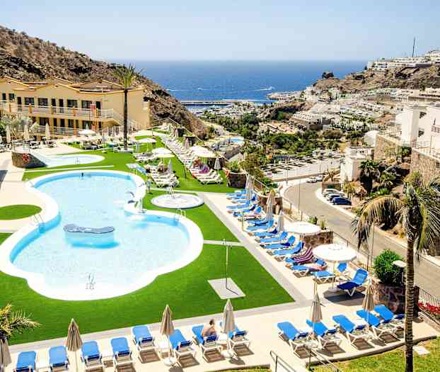 Bassengområde med solsenger og parasoller, omgitt av bygninger og fjell, med havet i bakgrunnen. (AI-generert tekst)