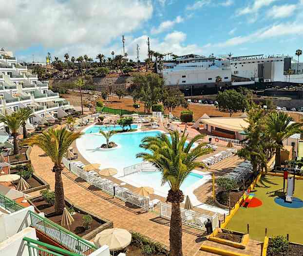 Bilde av et feriested med et basseng omgitt av solsenger og parasoller. I forgrunnen sees palmer og en lekeplass for barn. (AI-generert tekst)