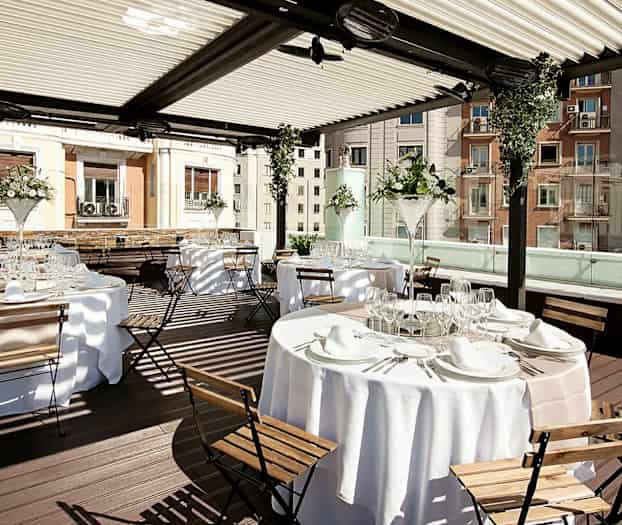 Bildet viser et elegant utendørsområde med dekkede bord og trestoler under en overbygd pergola. (AI-generert tekst)