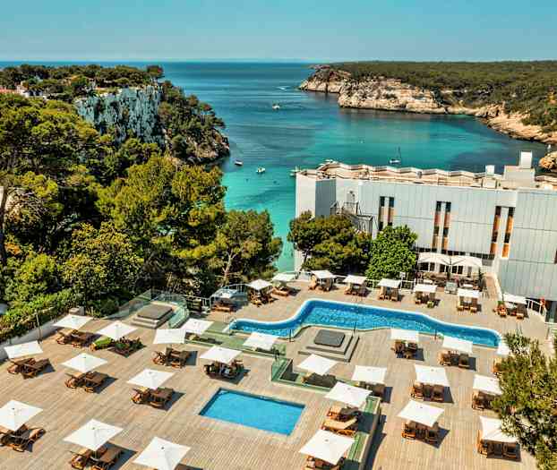 Et hotellområde med bassenger og solsenger ligger ved en bukt med klart vann og klippekyst. (AI-generert tekst)