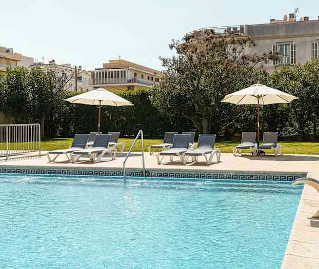 Et utendørs svømmebasseng med blått vann har solsenger og parasoller på et flislagt område ved siden av plen og grønne busker. (AI-generert tekst)