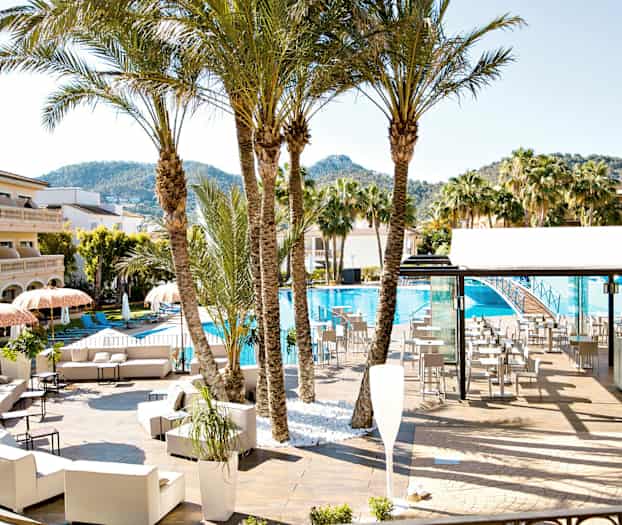 Et bassengområde med solsenger, parasoller, palmer og en bygning i bakgrunnen. (AI-generert tekst)