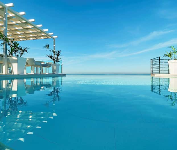 Et infinitybasseng med utsikt over havet, med bord og stoler under en skyggefull struktur ved siden av bassenget. (AI-generert tekst)