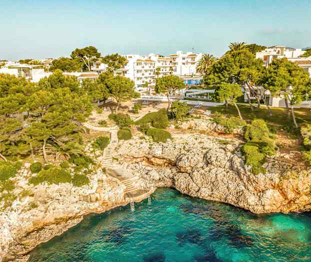 En kystlinje med klart blått vann, klipper samt trær og bygninger i bakgrunnen. (AI-generert tekst)