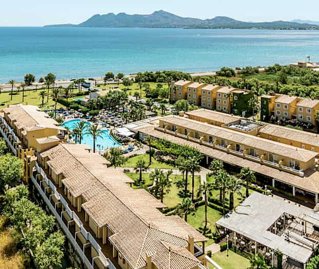 Flyfoto av et feriekompleks ved sjøen med basseng, grøntområder og flerboligbygninger. (AI-generert tekst)