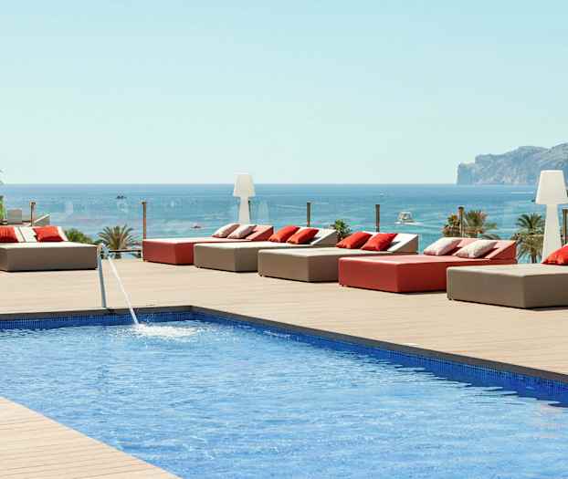 Utsikt over et basseng med solsenger og puter på en treterrasse, med havet og et fjell i bakgrunnen. (AI-generert tekst)