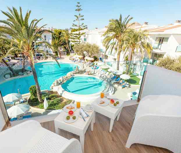 Utsikt fra terrasse mot bassengområde med solsenger, palmer og frokostbord. (AI-generert tekst)