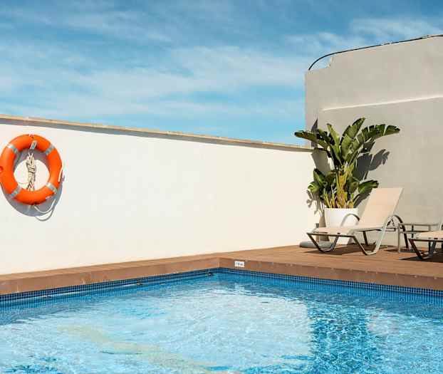 Et utendørs svømmebasseng med to solsenger og en oransje livbøye hengende på en hvit vegg. (AI-generert tekst)